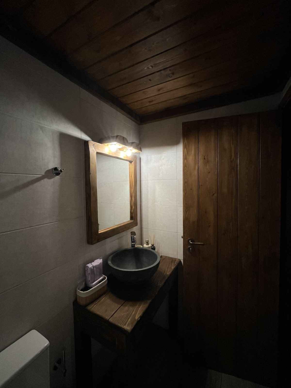 A compact bathroom is featured, showcasing a wooden sink vanity with a round basin. A framed mirror hangs above the sink, illuminated by warm light fixtures. The wood-paneled ceiling and natural wood door add a rustic touch to the design.
