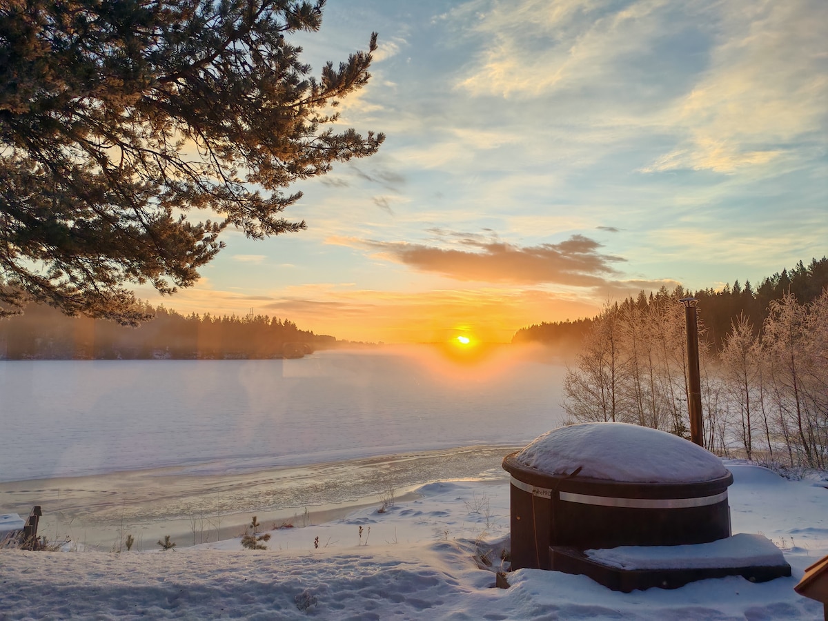 A scenic view of a river at sunrise, surrounded by snow and forest. The image features a hot tub in the foreground, offering a serene place to relax. Soft hues of orange and yellow illuminate the sky, blending with cool tones of the snowy landscape.