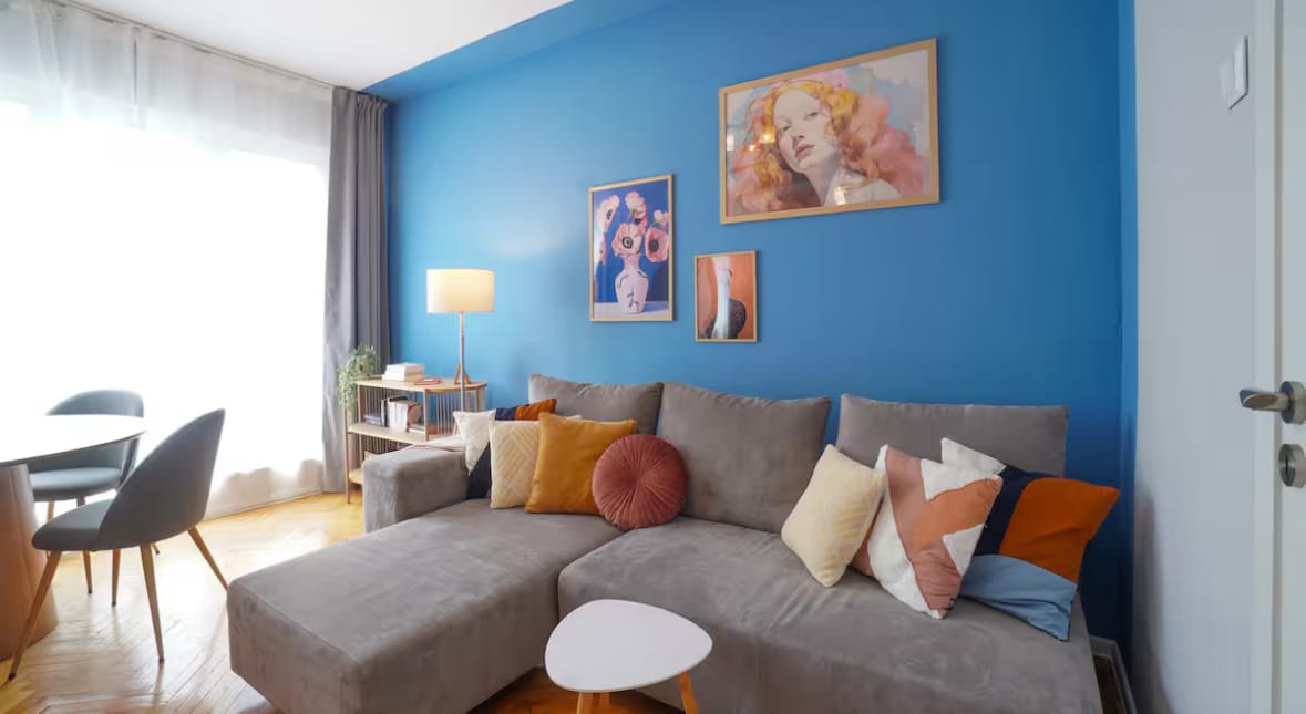 A modern living area features a spacious gray sectional sofa adorned with colorful pillows. A side table is positioned nearby, complementing the blue accent wall. Natural light floods the space through sheer curtains, highlighting the contemporary decor and nearby dining area.