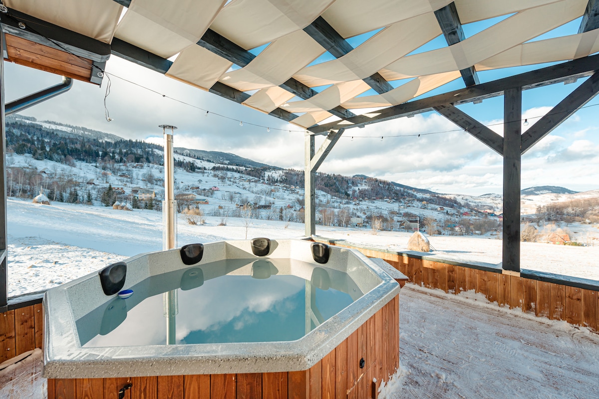 A covered jacuzzi area is presented, featuring a circular hot tub surrounded by a wooden deck. The serene winter landscape is visible in the background, with rolling hills and patches of snow, providing a tranquil setting for relaxation.