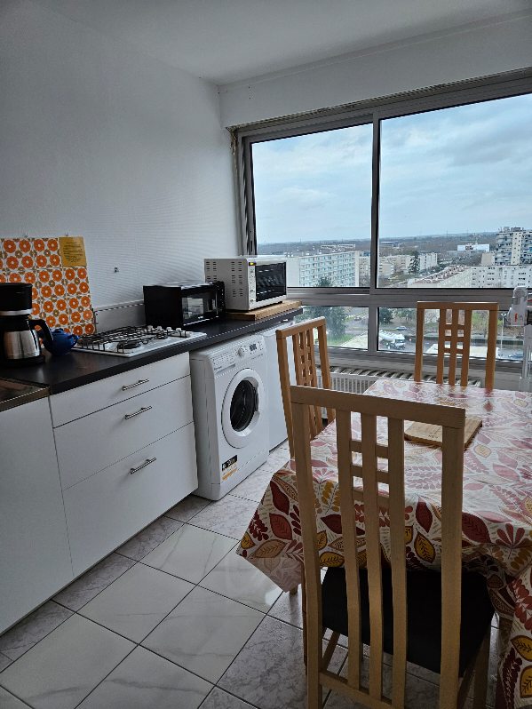 A well-equipped kitchen area features a washing machine, coffee maker, and microwave. The dining table, set with a patterned tablecloth, is surrounded by tall wooden chairs. Large windows provide a panoramic view of the neighborhood, allowing natural light to fill the space.