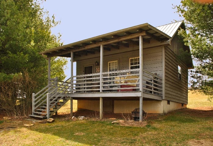 A wooden cabin is elevated on a foundation, featuring a covered porch with a railing. Sturdy steps lead to the entrance, which is flanked by large windows. Surrounding greenery and a natural landscape provide a serene backdrop, enhancing the cabin's secluded charm.