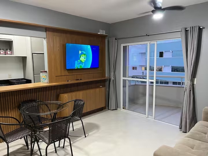 A modern living area features a sleek kitchen with wooden cabinetry and a compact dining set. A flat-screen TV is mounted on the wall, while large glass doors open to a balcony. Neutral tones and soft lighting create a relaxed environment.