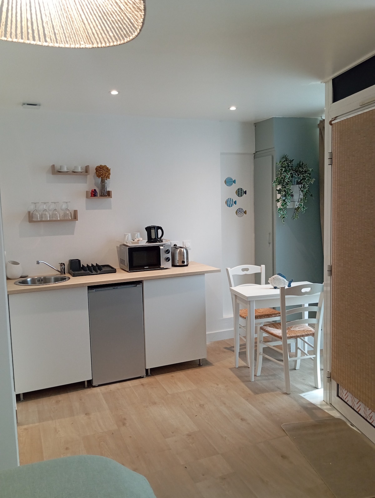 A kitchenette area is presented, equipped with essential appliances including a small refrigerator, microwave, and coffee maker. Light wooden flooring complements the white cabinetry. A small dining table with two chairs is included, adding functionality to this bright and welcoming space.