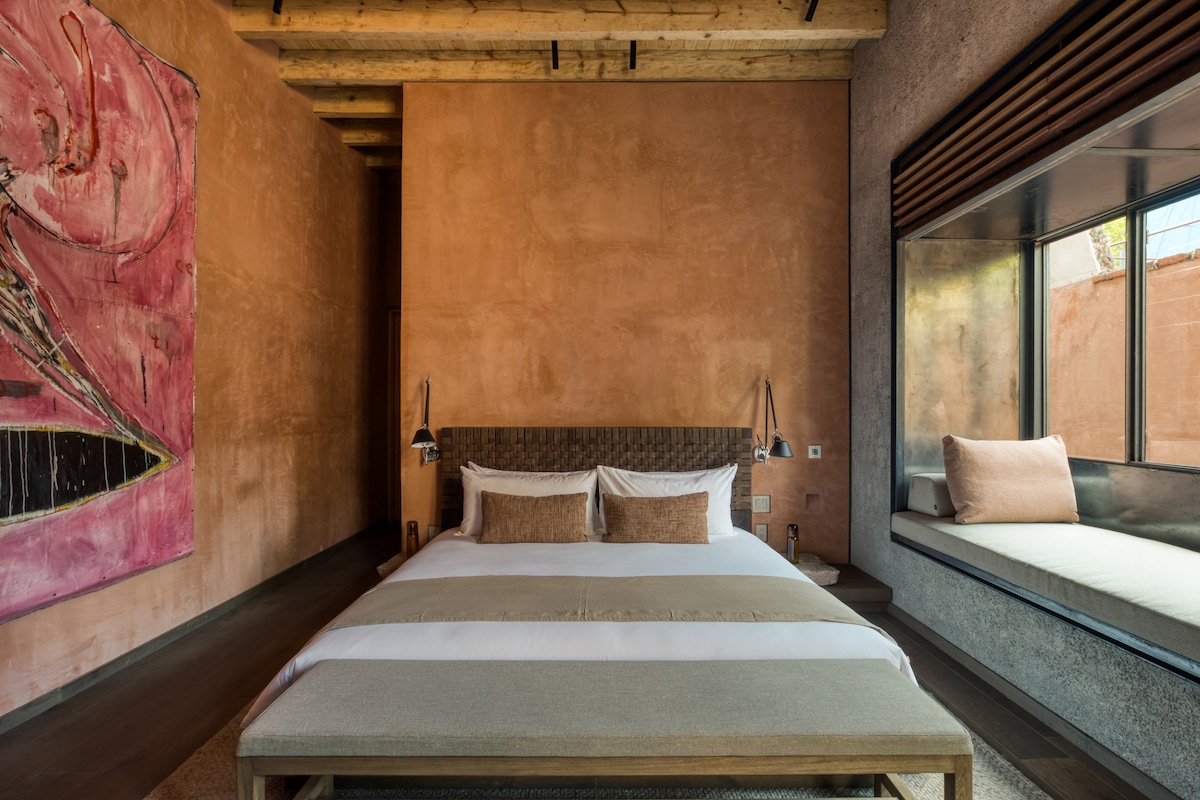 A spacious sleeping area features a large bed with neutral-toned bedding. Art with bold colors is displayed on the wall, complemented by wooden ceiling beams. A cozy seating nook is situated beside a window, allowing natural light to enter the room.