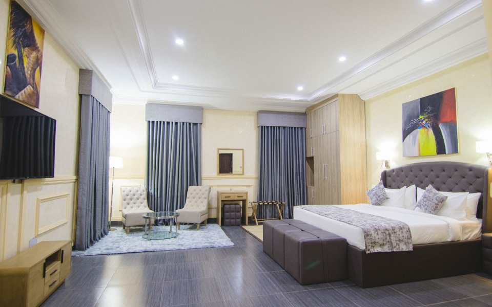 A spacious room features a large bed with a tufted headboard, complemented by contemporary artwork on the walls. Two armchairs and a small table are positioned near the window, with elegant drapes framing the view. A stylish cabinet and flat-screen television are included.