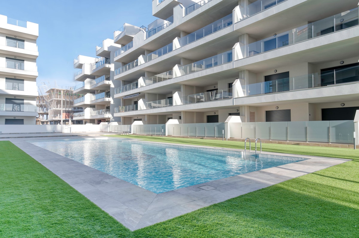 A communal swimming pool is surrounded by well-maintained grass lawns and modern apartment buildings. The pool features a clear, inviting surface reflecting the blue sky. Balconies are visible on the apartment facades, offering outdoor spaces for residents.