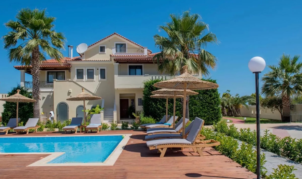 A charming villa is showcased with a clear blue sky above. Sun loungers with umbrellas surround a refreshing pool, complemented by landscaped gardens and palm trees, creating a relaxing outdoor atmosphere. The villa's architecture features a combination of light-colored walls and a terracotta roof.
