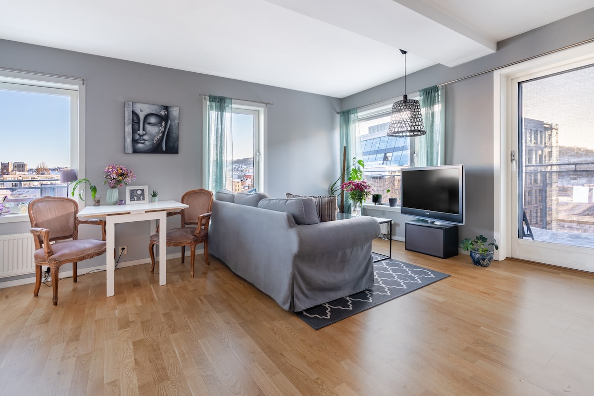 A modern living area features a comfortable gray sofa paired with two chairs at a small dining table. Natural light fills the space through large windows, illuminating wooden floors and contemporary decor. A television is positioned on a media unit, with access to a balcony visible.