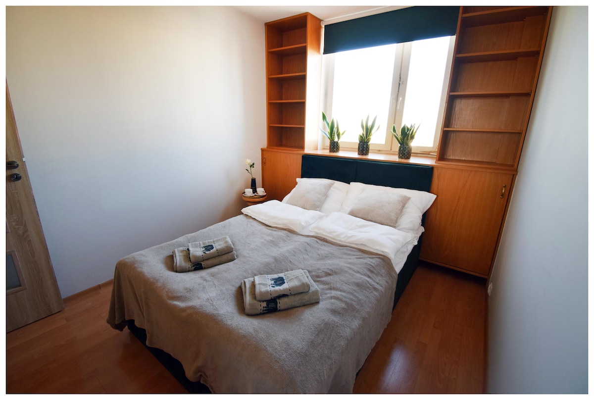 A cozy bedroom features a spacious bed with gently arranged white linens and towels. Shelving units are positioned above the bed, adorned with potted plants. Natural light enters through large windows, enhancing the soft colors of the room.