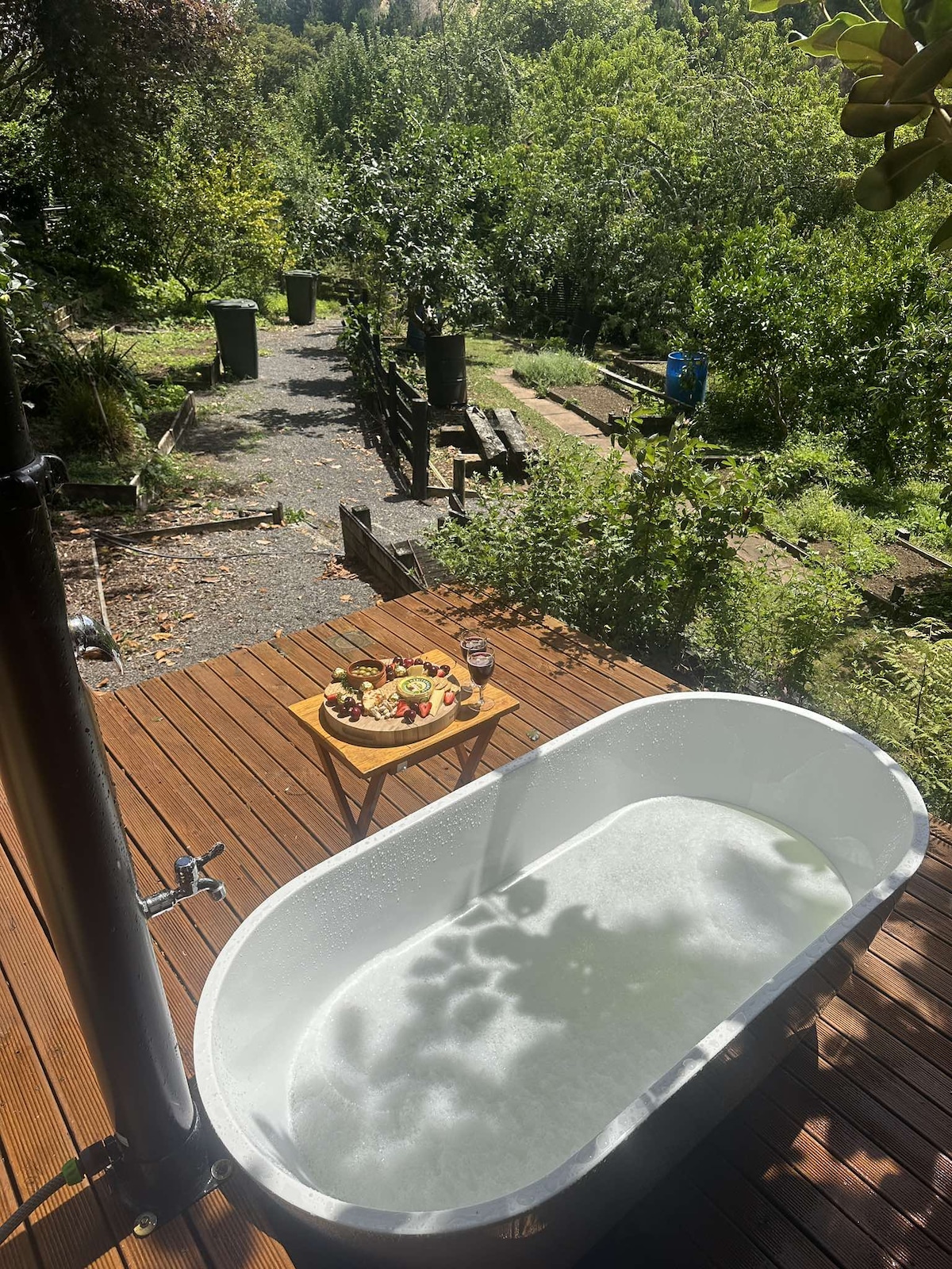 An outdoor tub sits on a wooden deck, surrounded by lush greenery. A small wooden table holds a platter of fruit and a glass, complementing the serene setting. Sunlight filters through the trees, creating gentle shadows on the tub's surface.