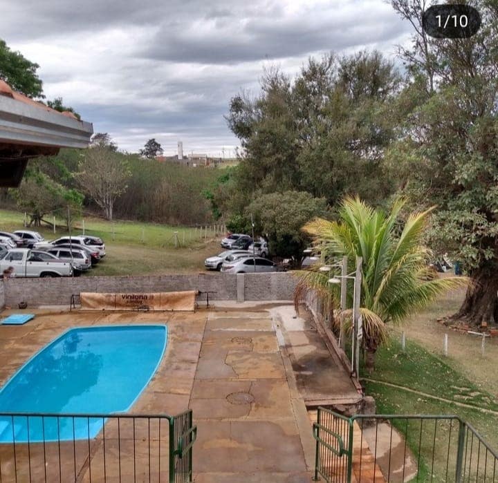 A pool area features a turquoise water surface surrounded by a stone deck. Lush greenery and trees are seen off to the side, along with a parking space accommodating multiple vehicles. A gentle overcast sky completes the outdoor view.