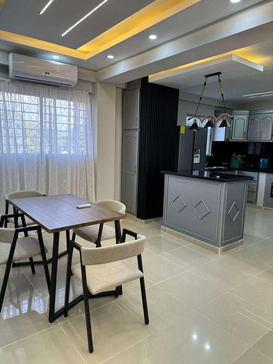 A dining area features a wooden table surrounded by four light-colored chairs. Natural light enters through sheer curtains, illuminating the open space that connects to a modern kitchen with sleek cabinetry and a central island accentuated by overhead lighting.