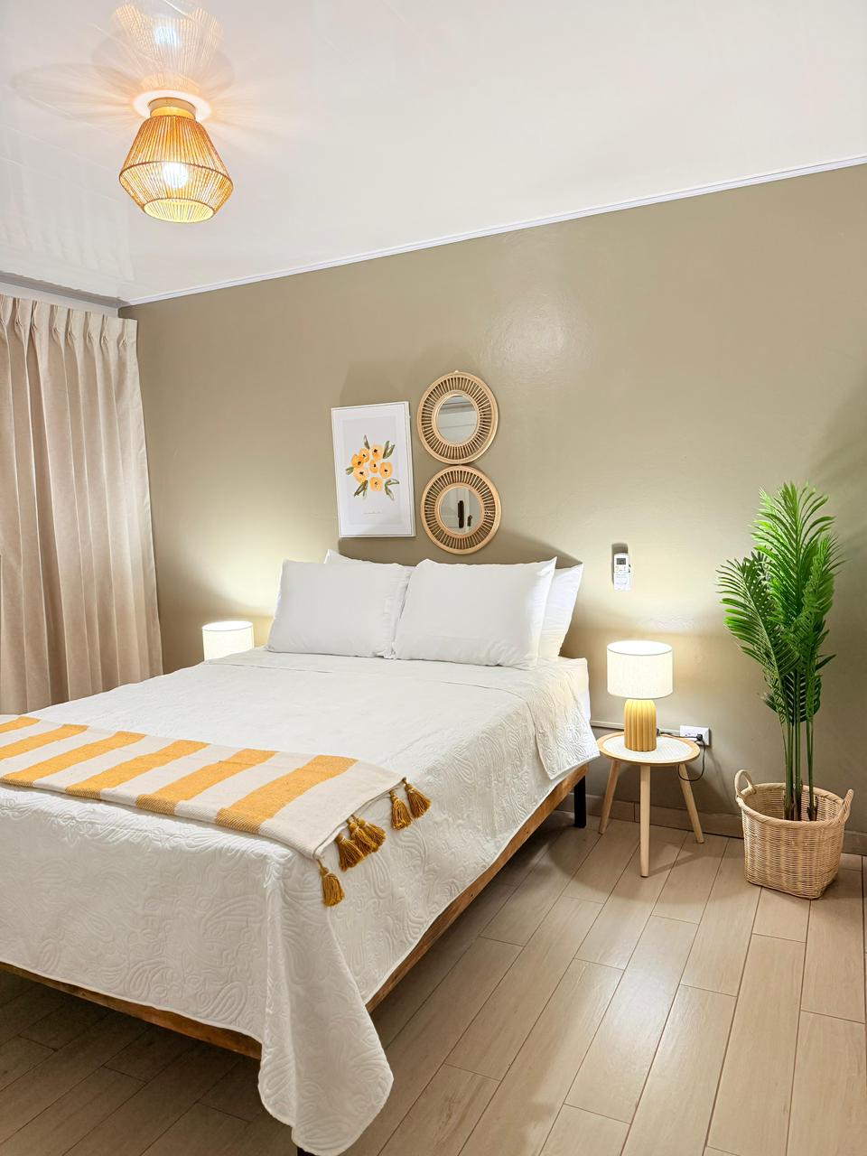 A well-lit sleeping area features a spacious bed adorned with white linens and a decorative blanket with yellow accents. Two bedside lamps provide soft lighting, while a potted plant adds a touch of greenery. The walls are painted in a calming neutral tone.