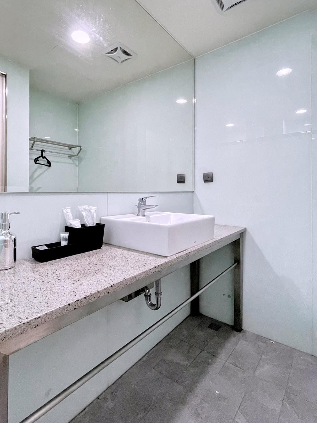 A modern bathroom is presented with sleek surfaces and ample lighting. A rectangular sink is positioned on a granite countertop, accompanied by a dispenser and neatly arranged toiletries. The walls are finished in smooth glass, enhancing the bright and clean ambiance.