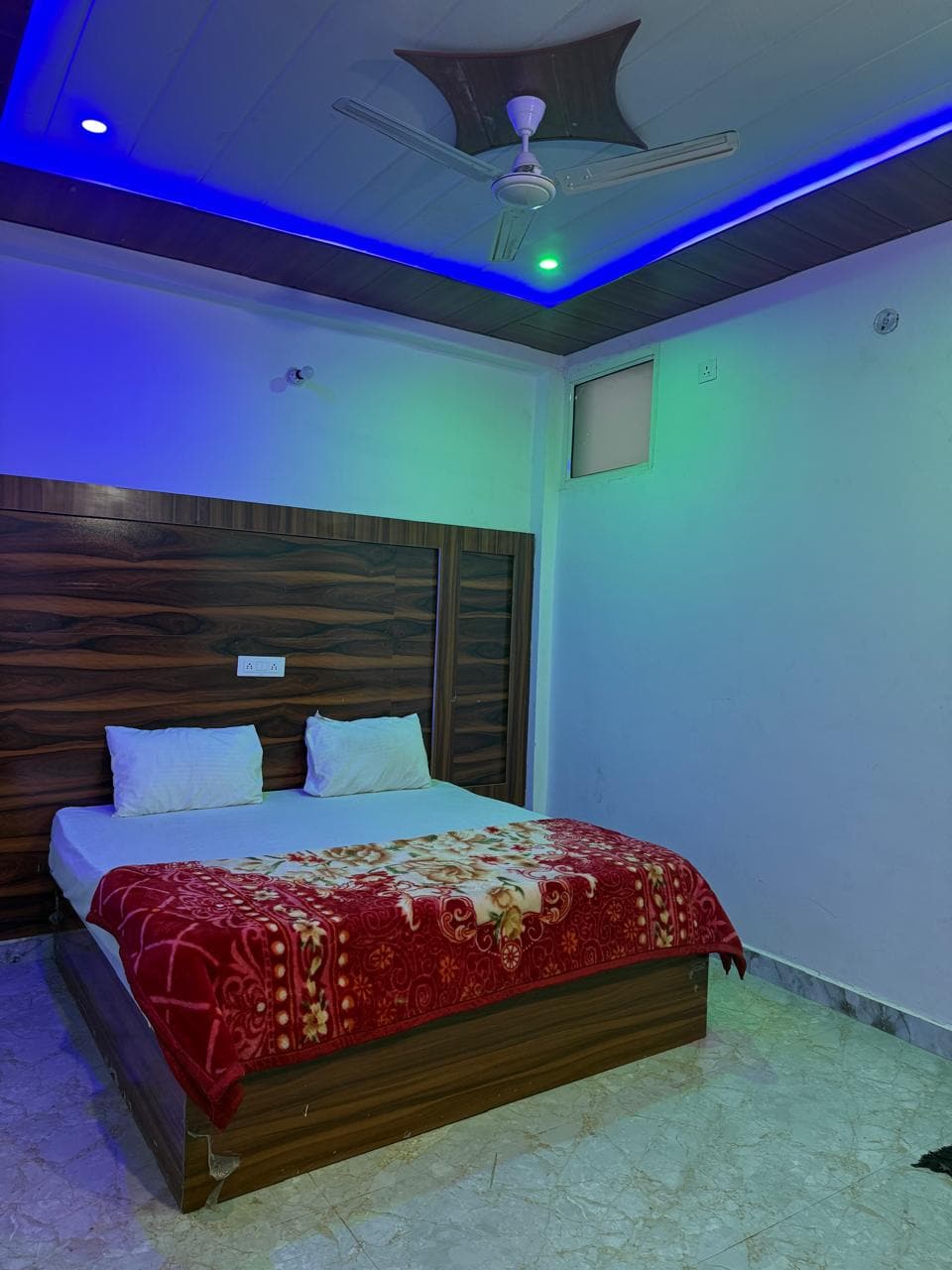 A bedroom is showcased with a spacious bed covered by a bright red blanket. Soft lighting with a bluish hue is emitted from the ceiling, highlighting the wooden paneling behind the bed. A ceiling fan and a window contribute to the overall layout of the room.
