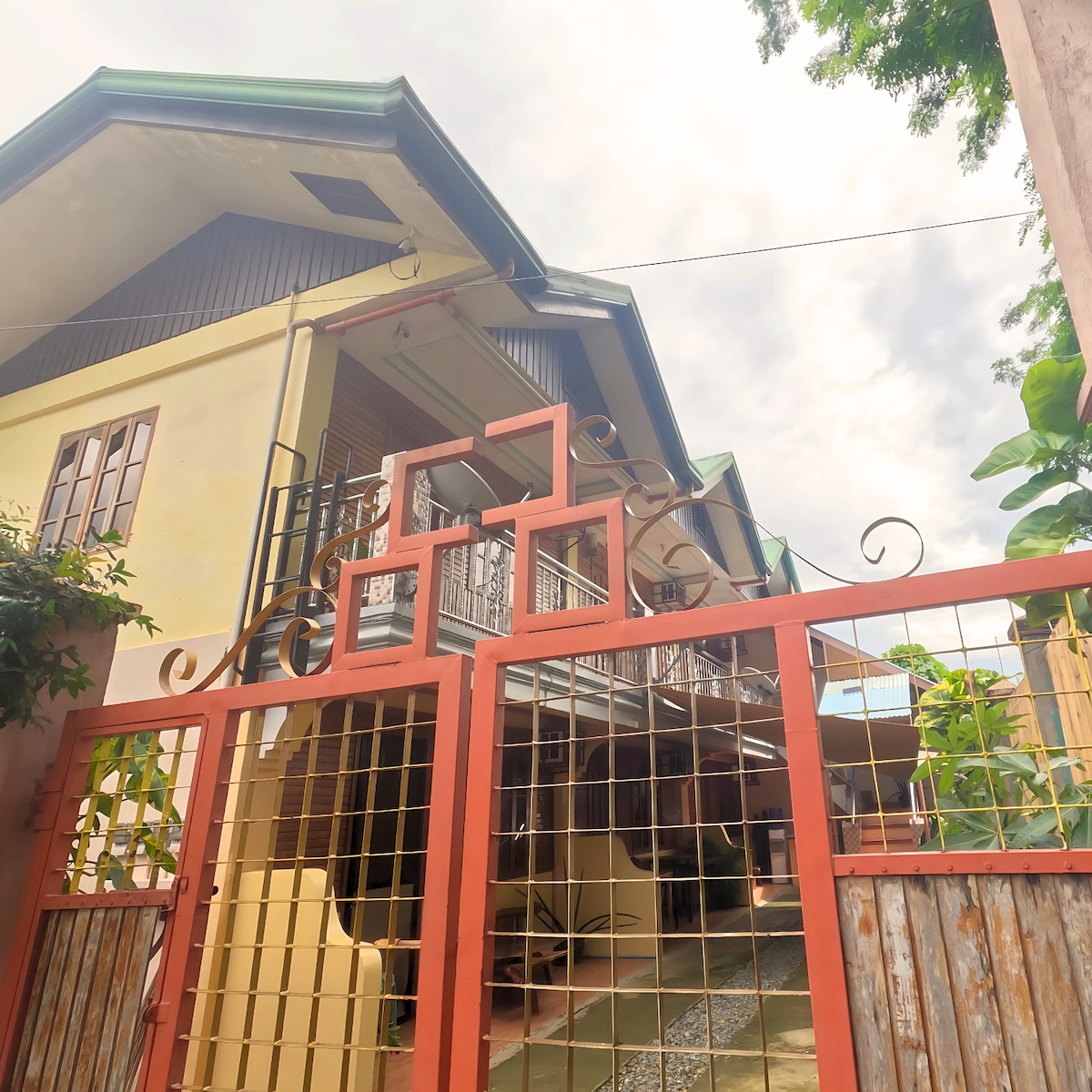 A welcoming entrance to the property is highlighted by a vibrant red gate, leading to a spacious patio area. The two-story building exhibits a mix of architectural styles, featuring balconies and large windows under a gently slanted roof. Lush greenery complements the entrance.