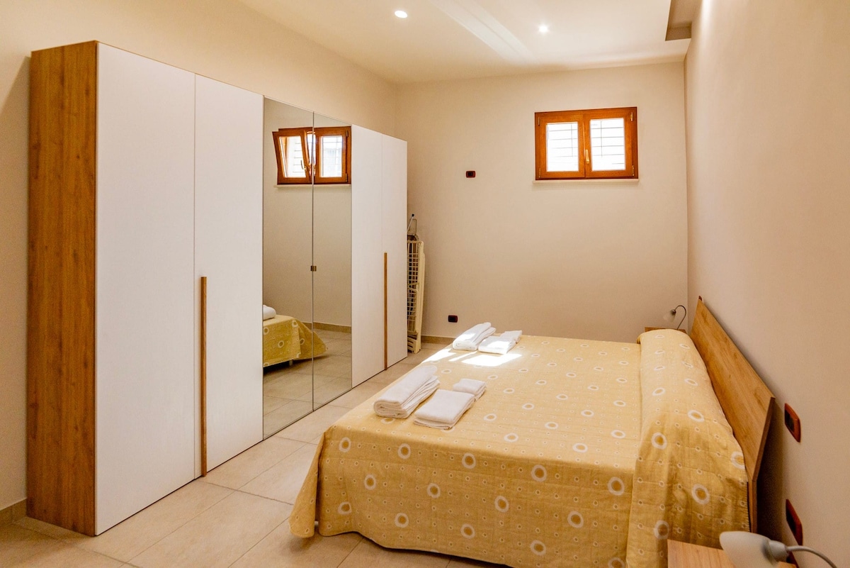 A spacious bedroom features a bed covered with a yellow patterned blanket, complemented by neatly arranged towels. A mirrored wardrobe reflects the light from the windows, which are equipped with wooden shutters. Soft lighting enhances the room's calm and organized appearance.