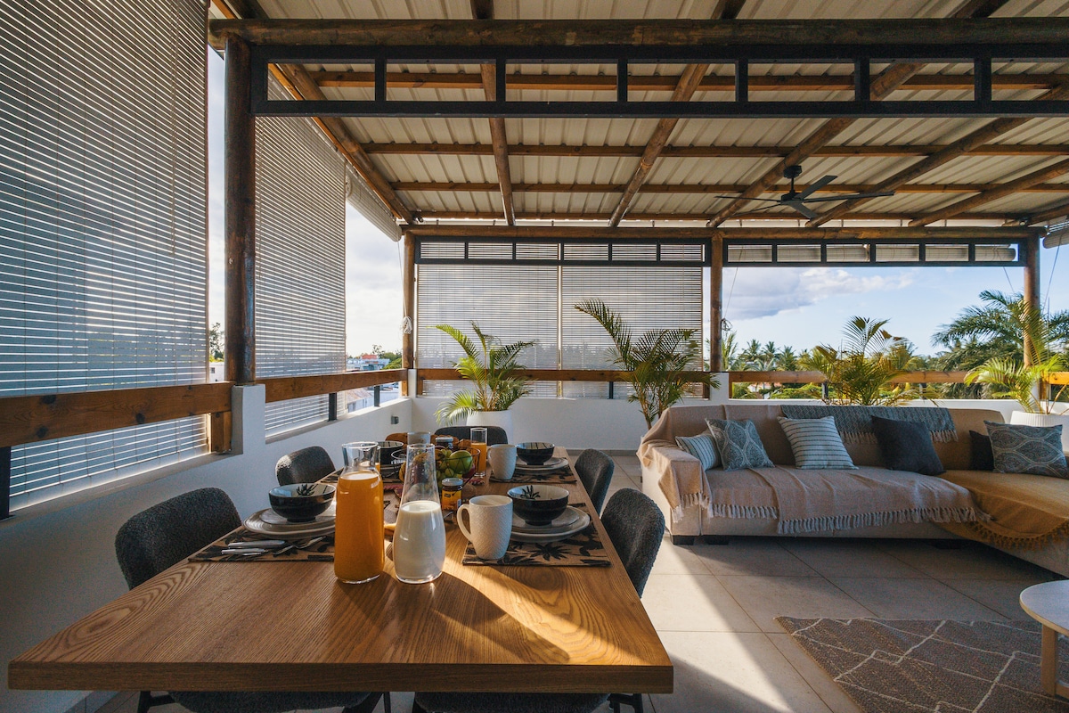A spacious terrace features a wooden dining table set for breakfast, complemented by a cozy seating area with light-colored cushions. Sunlight filters through the blinds, highlighting the greenery in the background and creating a relaxed atmosphere for outdoor dining.