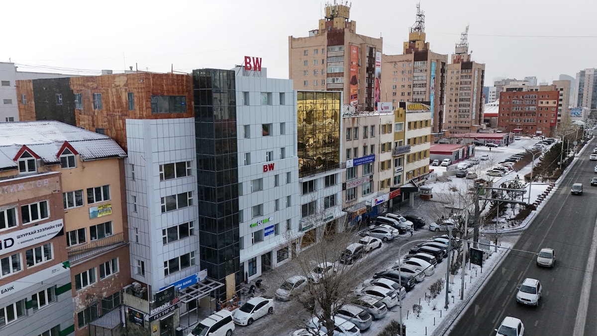 A cityscape is shown, featuring a mix of modern and traditional buildings along a snowy street. Several vehicles are parked along the roadside, while busy commercial establishments with signs in various languages are visible. The scene reflects an urban atmosphere, typical of a vibrant downtown area.
