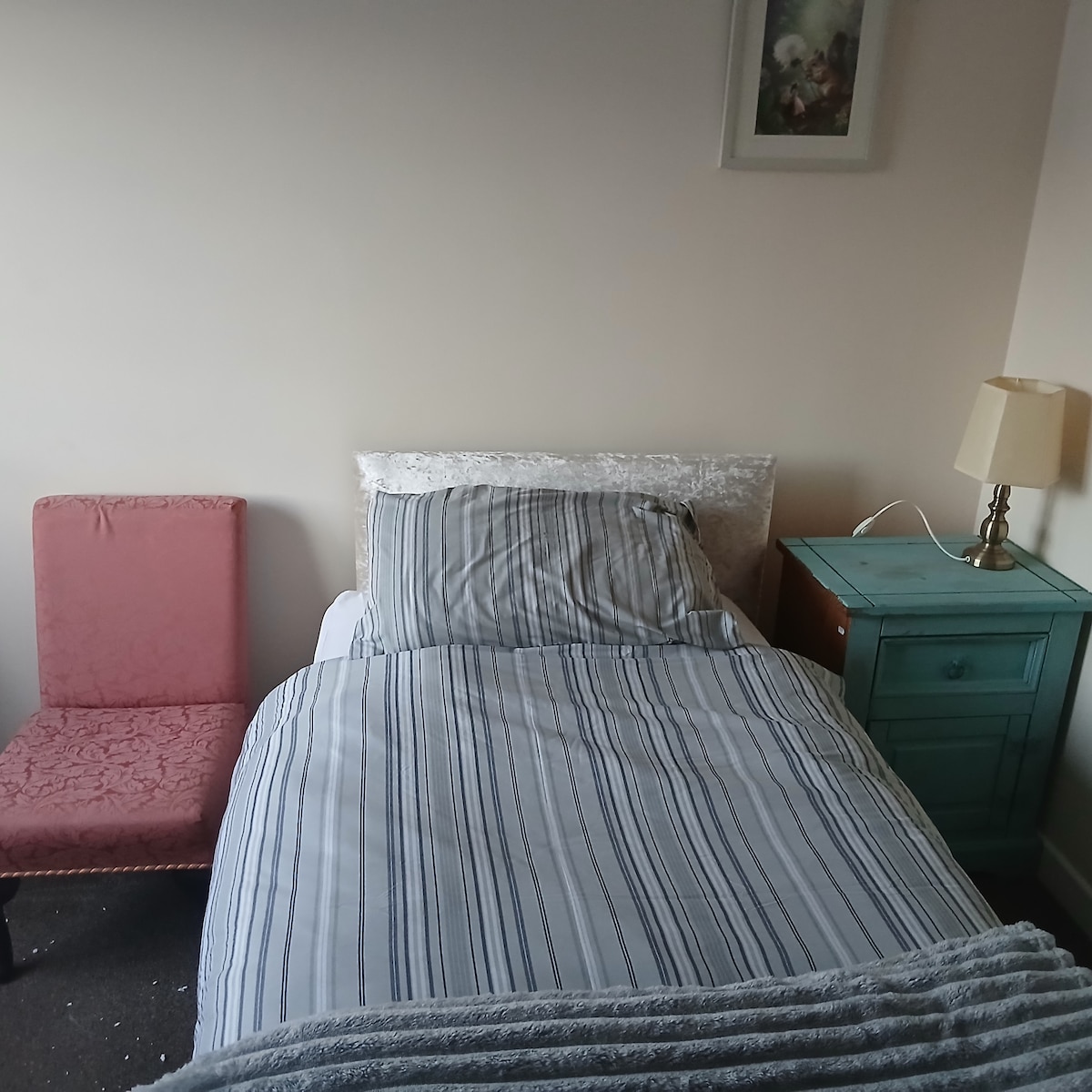 A cozy bedroom features a spacious bed dressed in striped bedding. A pink chair sits beside the bed, and a small turquoise nightstand holds a lamp. Light colors on the walls create a bright atmosphere, complemented by a framed artwork hanging above.