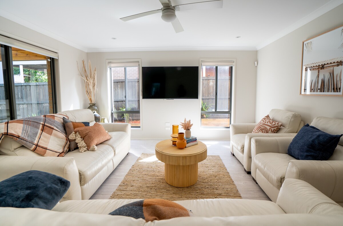 A light-filled living area features two cream sofas adorned with decorative pillows. A round wooden coffee table sits at the center, complemented by a woven rug. Large windows allow natural light to illuminate the space, accentuating a large flat-screen TV mounted on the wall.