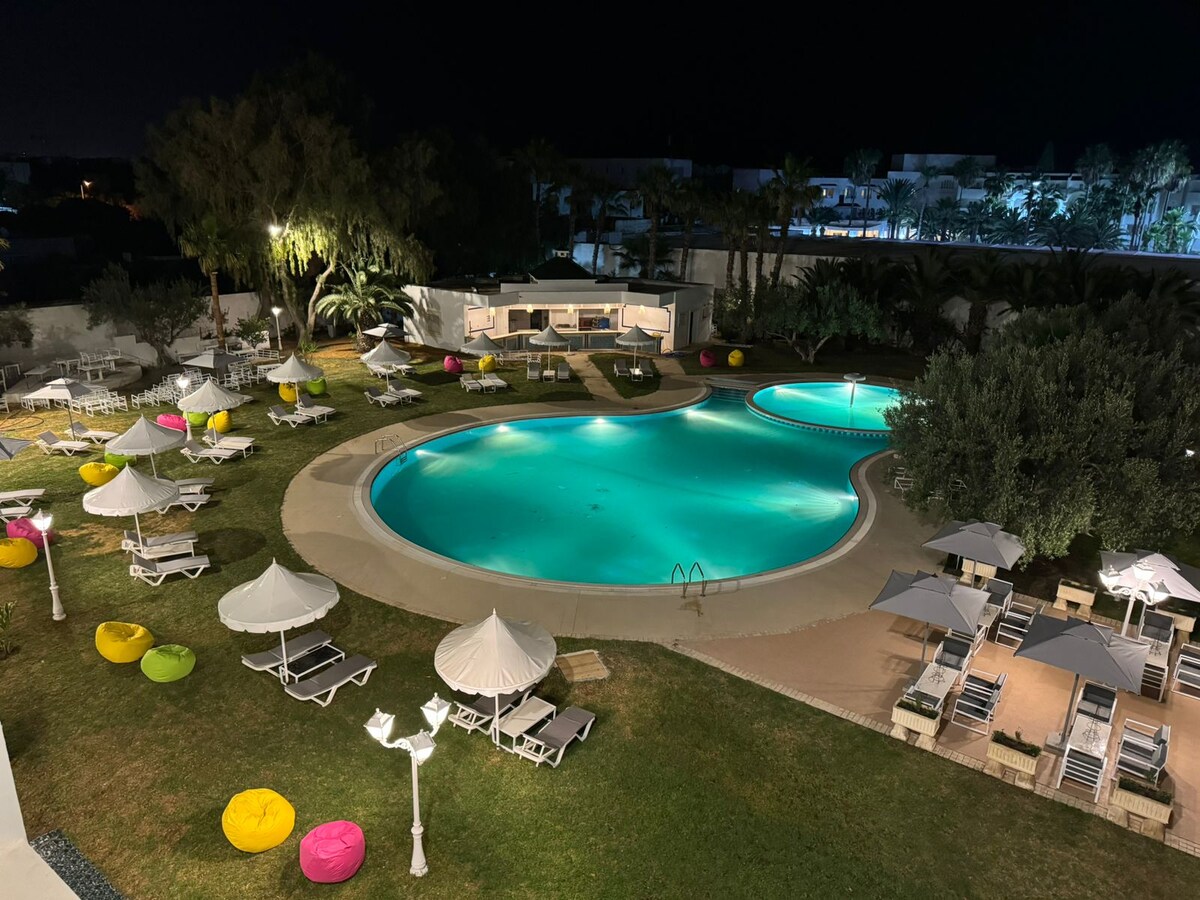 A large swimming pool is illuminated by soft lights, surrounded by well-maintained grass. Colorful lounge chairs and umbrellas are arranged around the pool. A seating area is visible nearby, adding to the relaxation options in this inviting outdoor space.