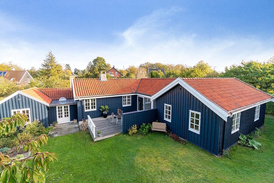 A charming summer house is presented in blue with a red-tiled roof, surrounded by a well-maintained green lawn. Several windows allow natural light into the interior. A wooden bench sits on the patio, facilitating outdoor relaxation.