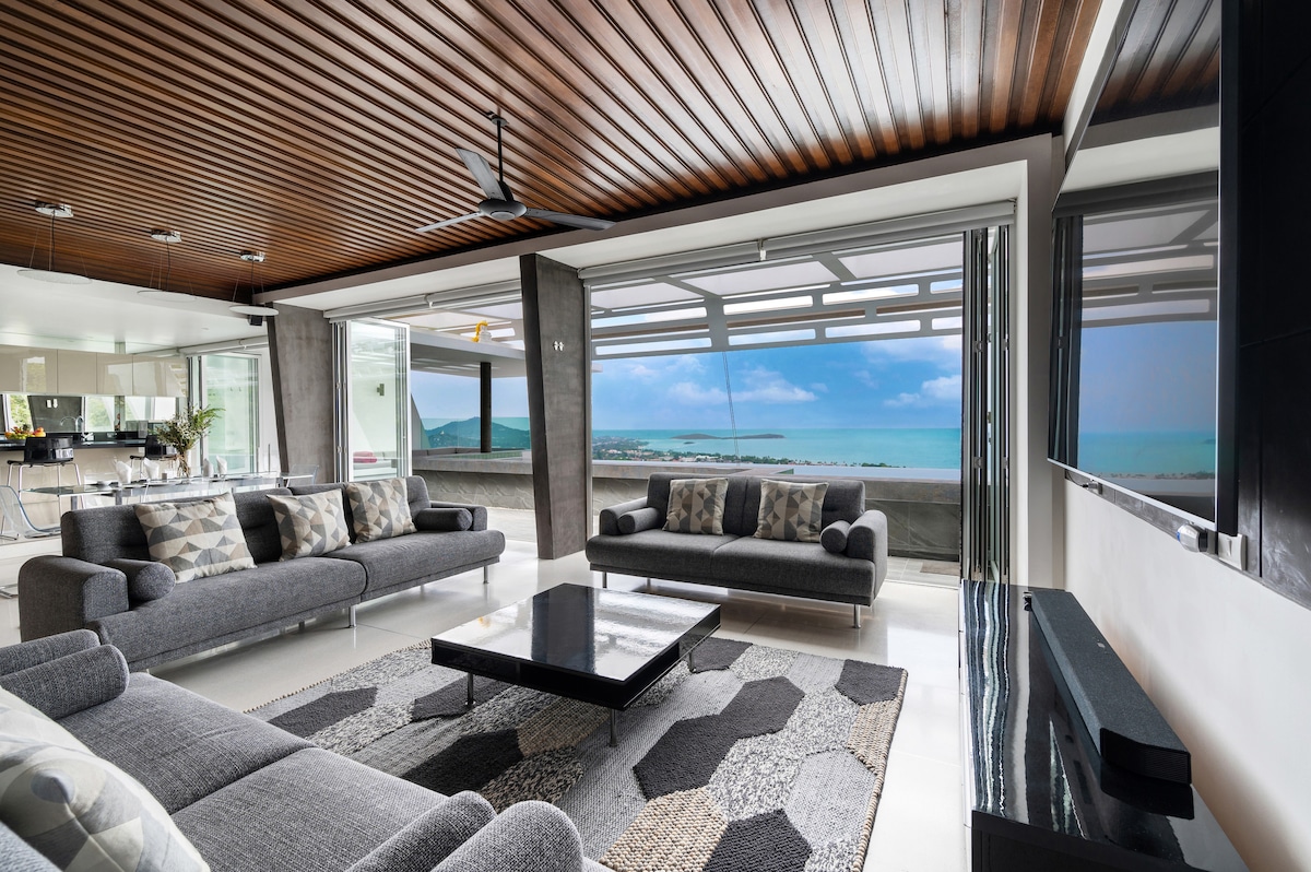A modern living area is displayed with large windows revealing expansive views of the ocean and surrounding hills. Comfortable gray sofas surround a dark coffee table, while a patterned area rug adds visual interest to the light-colored flooring. A sleek entertainment unit is positioned along one wall.