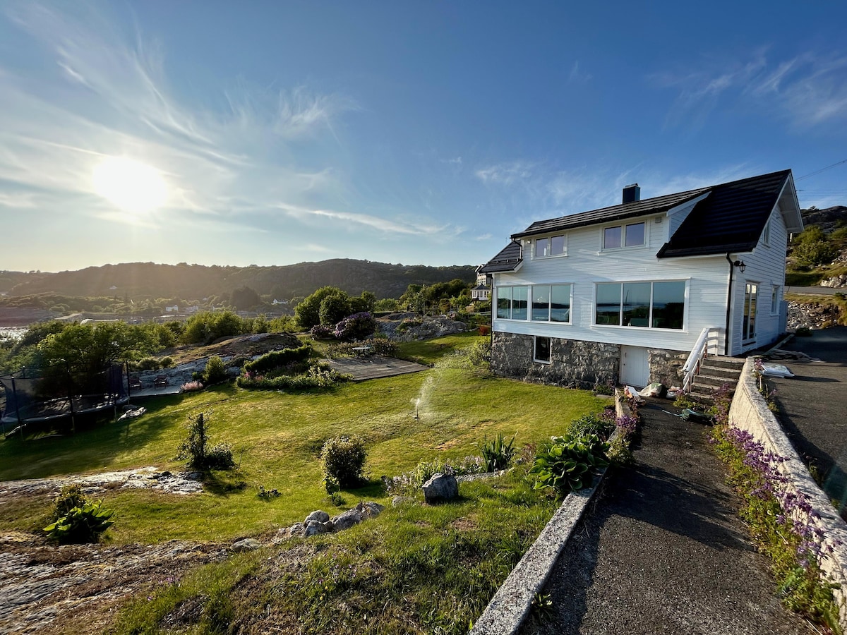 A large family house is set against a scenic backdrop, featuring expansive glass windows. A well-maintained garden surrounds the property, with various plants and greenery. A pathway leads to the entrance, while the sun sets in the background, illuminating the landscape.