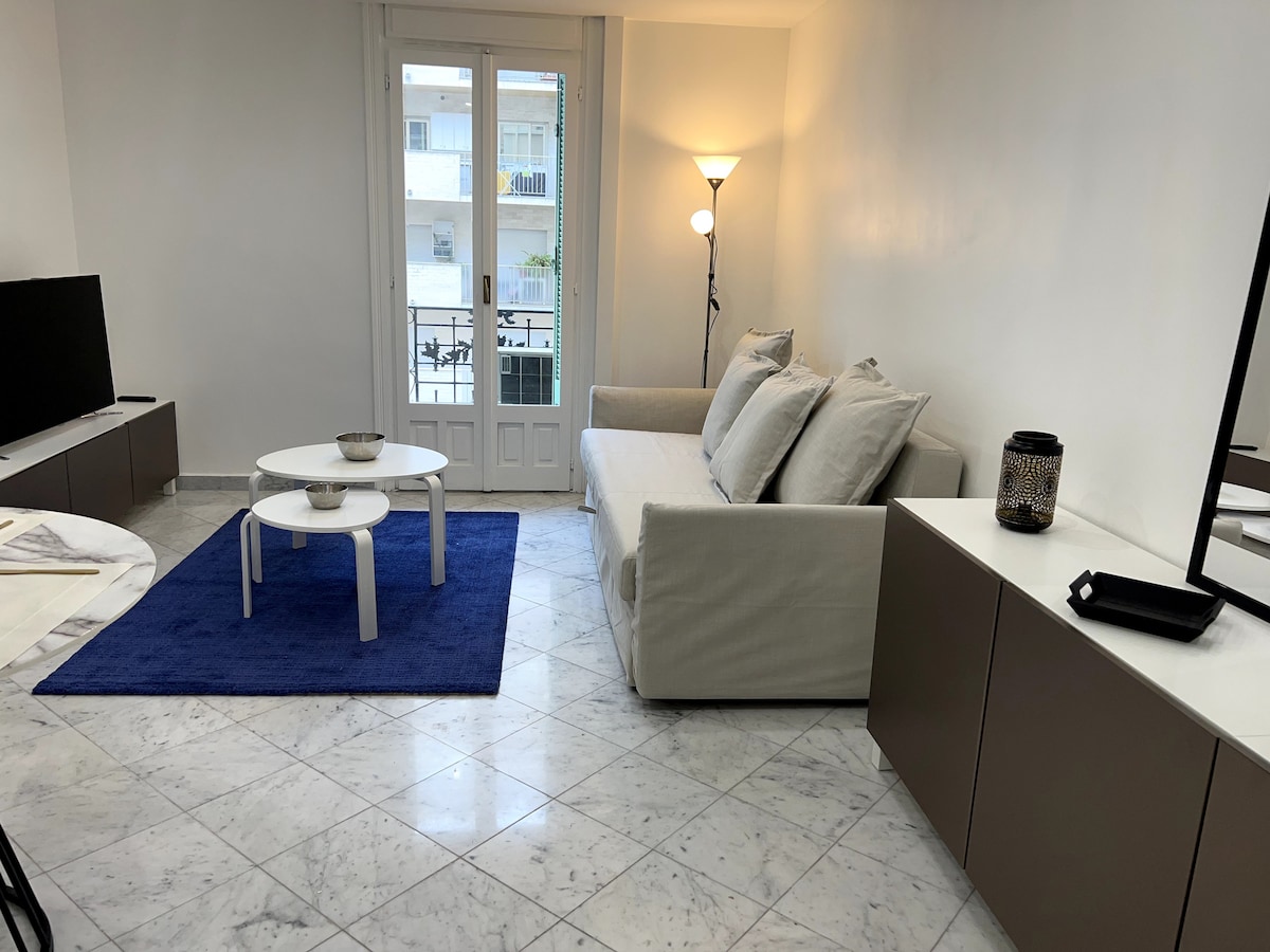 A bright living area features a light-colored sofa with soft cushions, accompanied by two round coffee tables set on a blue area rug. Natural light enters through glass doors, illuminating the marble flooring and simple decor.