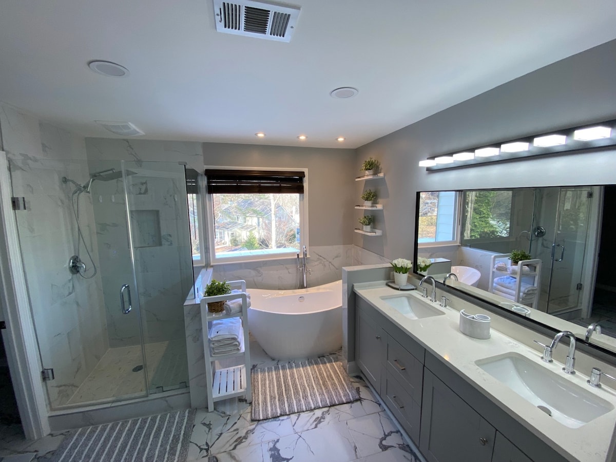 The spacious bathroom features a glass-enclosed shower and a freestanding soaking tub positioned near a window. Natural light illuminates the room, highlighting elegant marble tiles and a dual-sink vanity. Shelves with potted plants add a touch of greenery to the soothing decor.