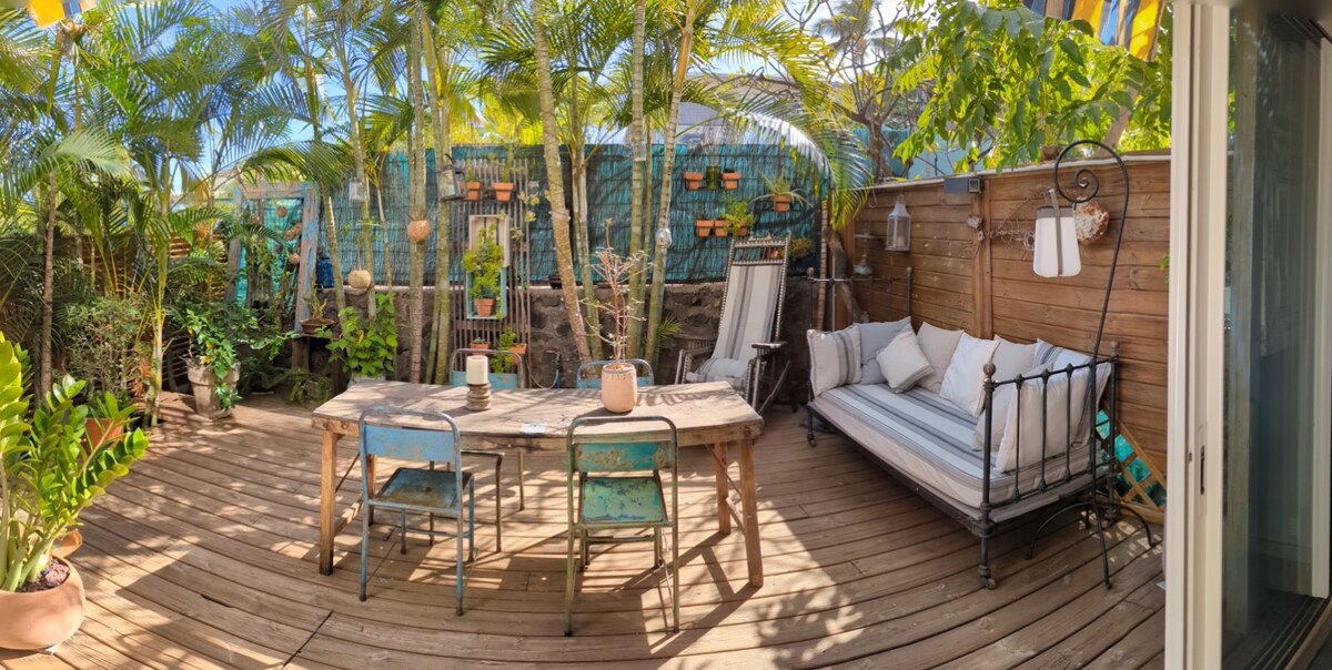 A wooden terrace is surrounded by lush greenery, featuring a large dining table accompanied by two metal chairs. A cozy seating area with a daybed is accented by white cushions. Hanging plants and decorative lanterns add a touch of charm to the outdoor space.