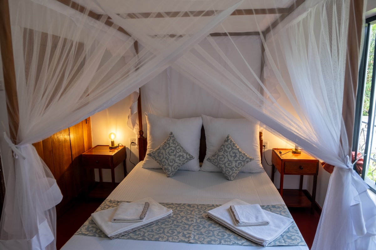 A cozy sleeping area is featured, showcasing a spacious bed draped with soft white mosquito netting. Two patterned pillows add a decorative touch. Bedside tables are located on either side, complemented by soft bedside lamps that provide warm lighting.