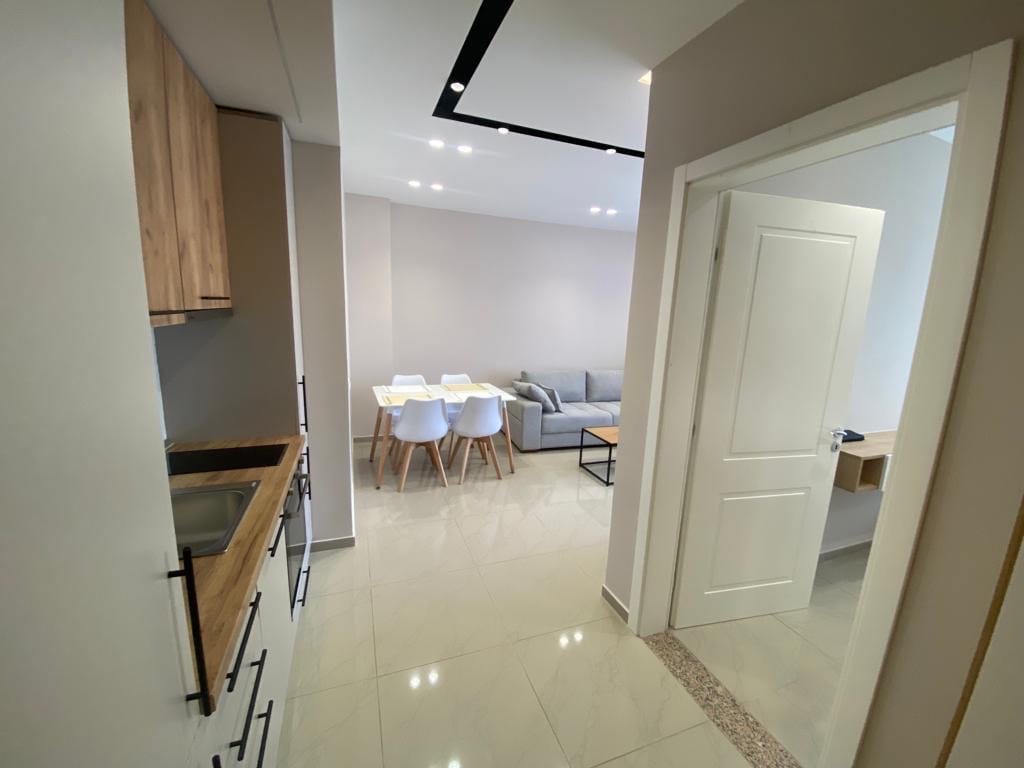 A modern hallway leads into a bright open living space. The kitchen features light wood cabinetry and a sink. The dining area includes a table with white chairs, while a spacious living room highlights a grey sofa, creating a welcoming environment for relaxation and gathering.