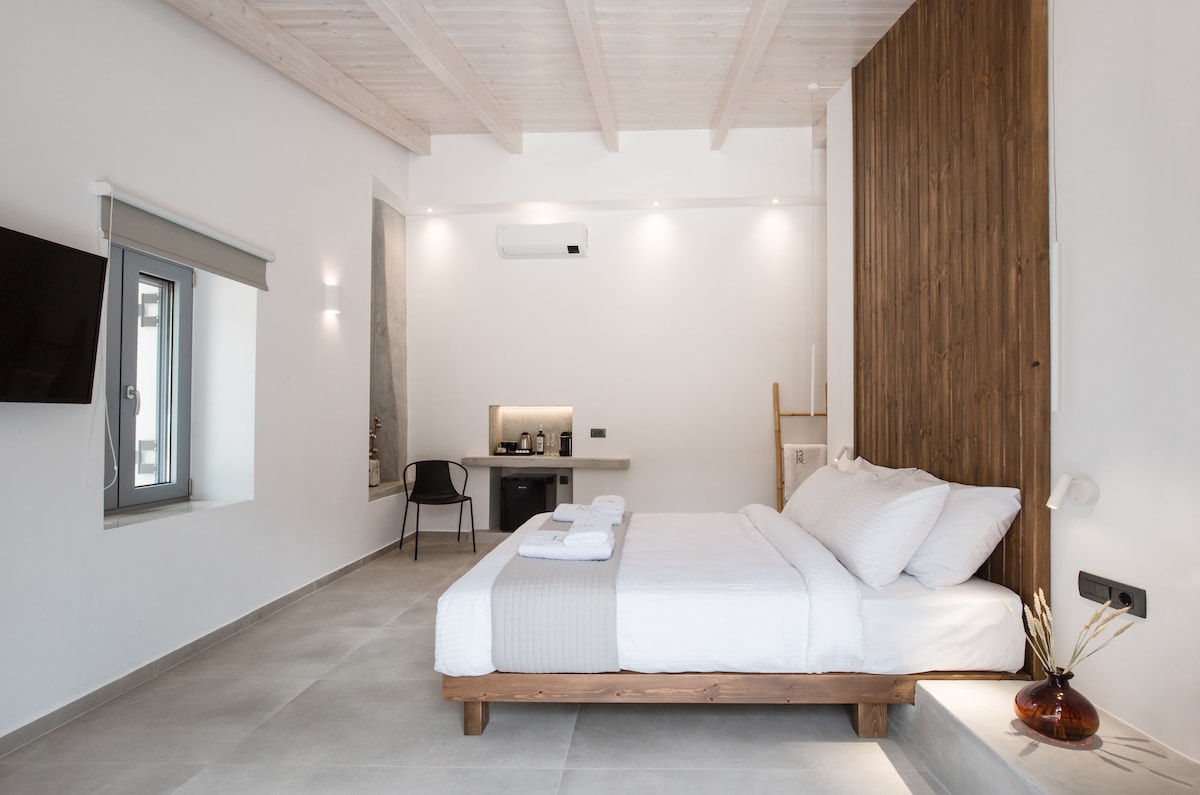 The cozy double room features a spacious bed with white linens against a wooden accent wall. A small work area with a chair and desk is visible on one side, while natural light enters through a window adorned with minimalistic treatments.
