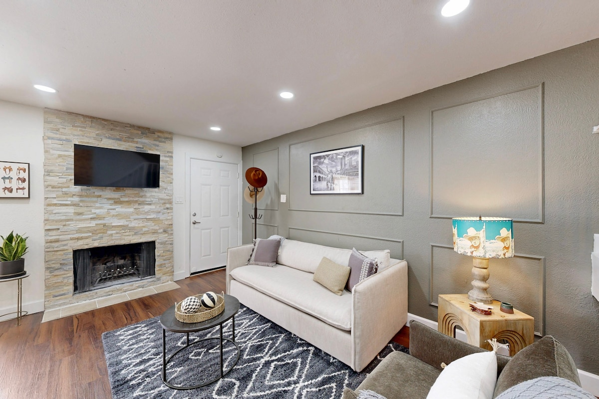 A cozy living room features a modern sofa facing a stylish stone fireplace. Wall-mounted artwork adds character, while a patterned rug complements the space. A decorative lamp and a small side table enhance the inviting atmosphere, with warm lighting illuminating the room.