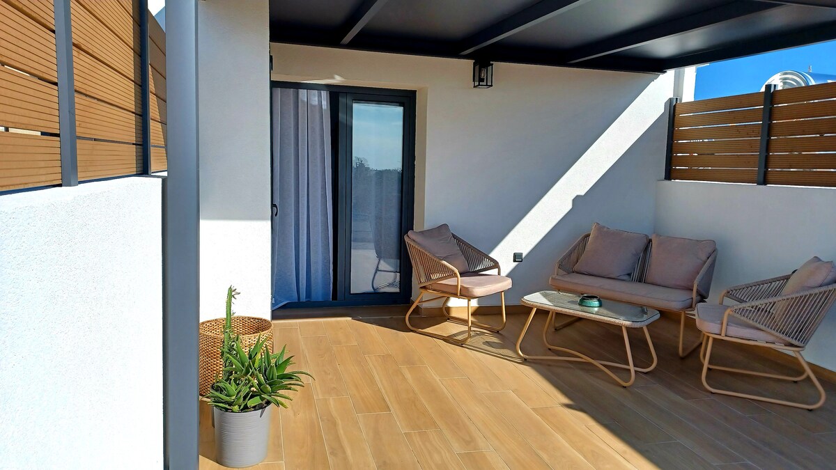 A private balcony offers a serene outdoor space with two comfortable chairs and a small coffee table. Sunlight filters through the protective slats, creating a warm atmosphere. A potted plant adds a touch of greenery against the light wooden flooring.