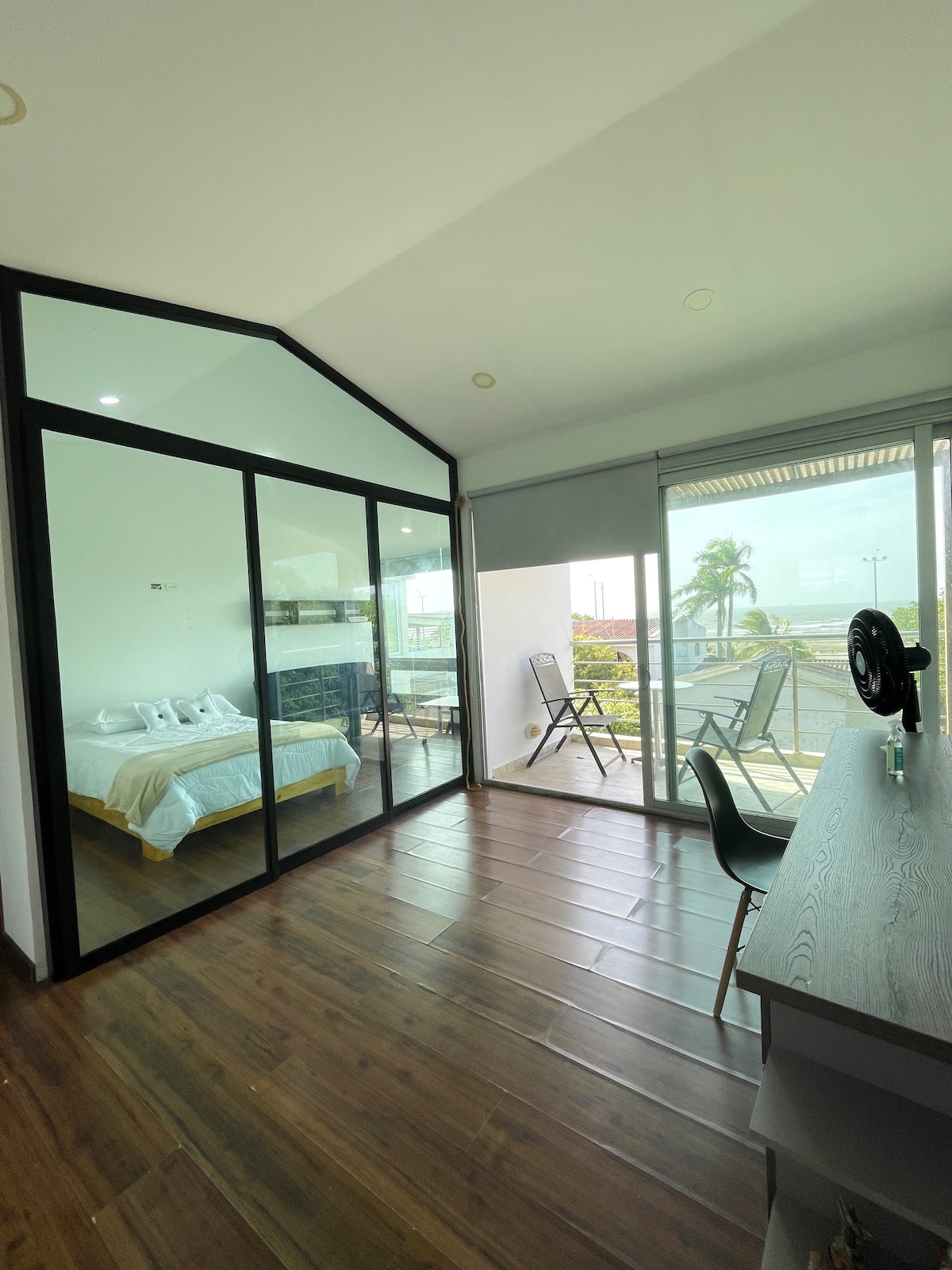 A bright loft space is showcased, featuring large glass doors that open to a private balcony. A bed with white linens is visible through a glass partition. Modern furnishings are found throughout, with a workspace that includes a desk and chair, enhancing the functional layout.