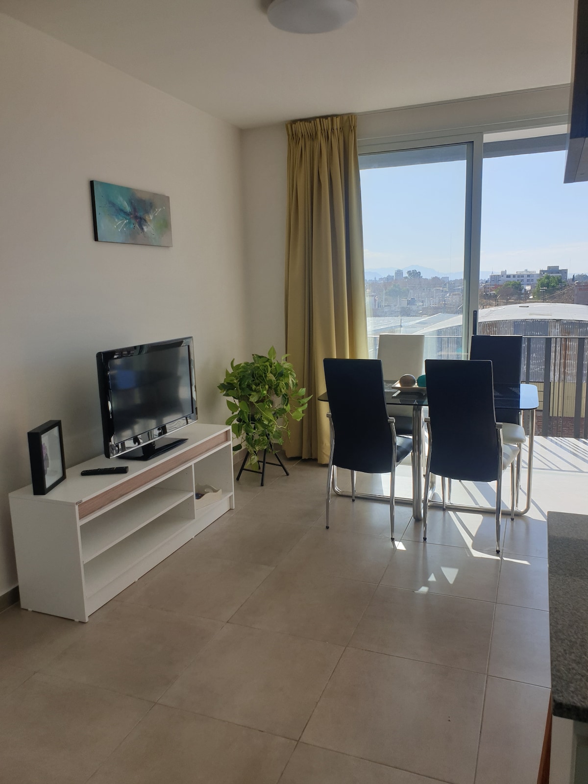 A bright living area features a flat-screen TV on a low white cabinet. A round dining table with four dark chairs is set near a large window, allowing natural light to flow in. A potted plant adds a touch of greenery to the space.