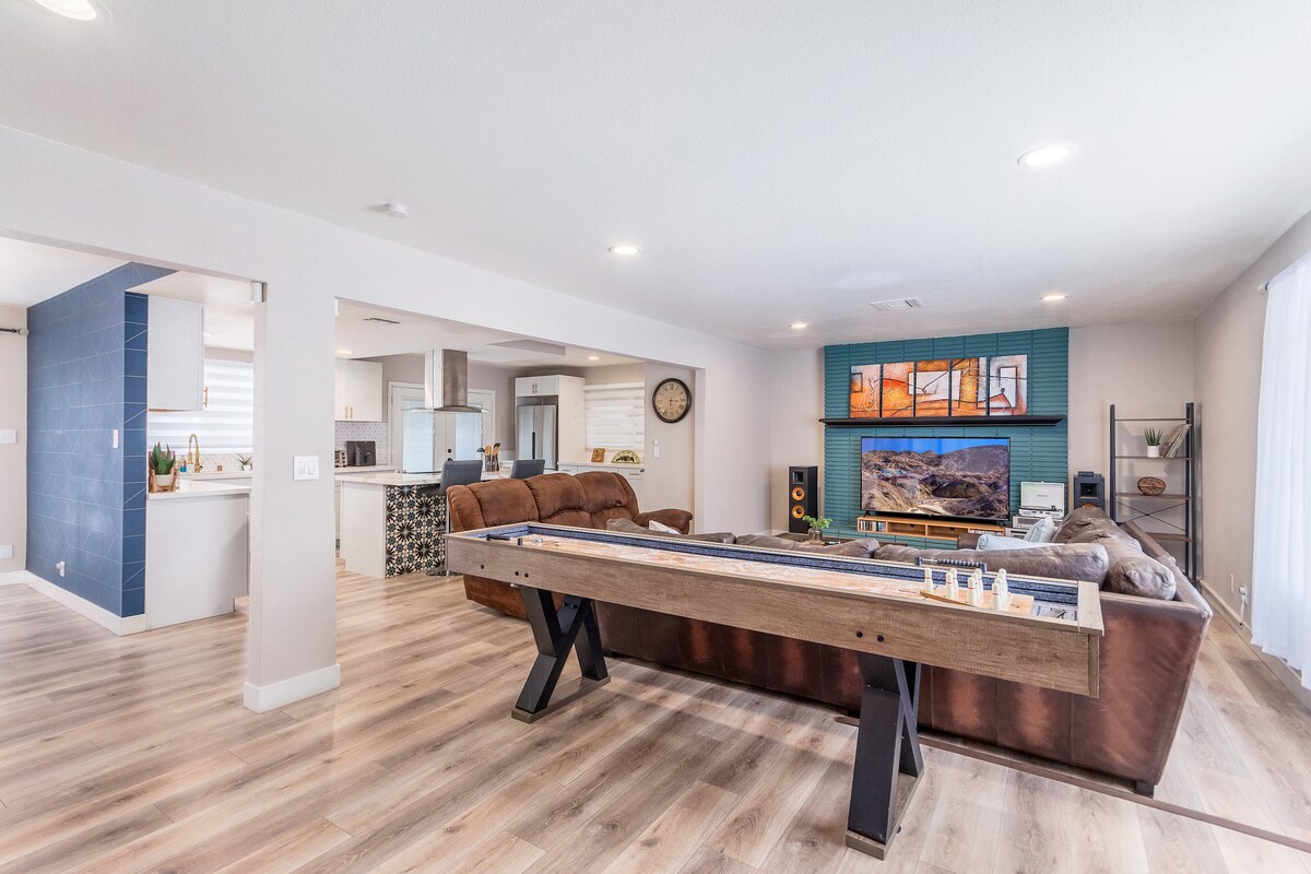 A spacious living area is showcased with a shuffleboard table and an inviting sofa set. A large television sits above a stylish media console beneath a colorful wall display. The open layout connects the kitchen area, featuring modern appliances and a sleek design.