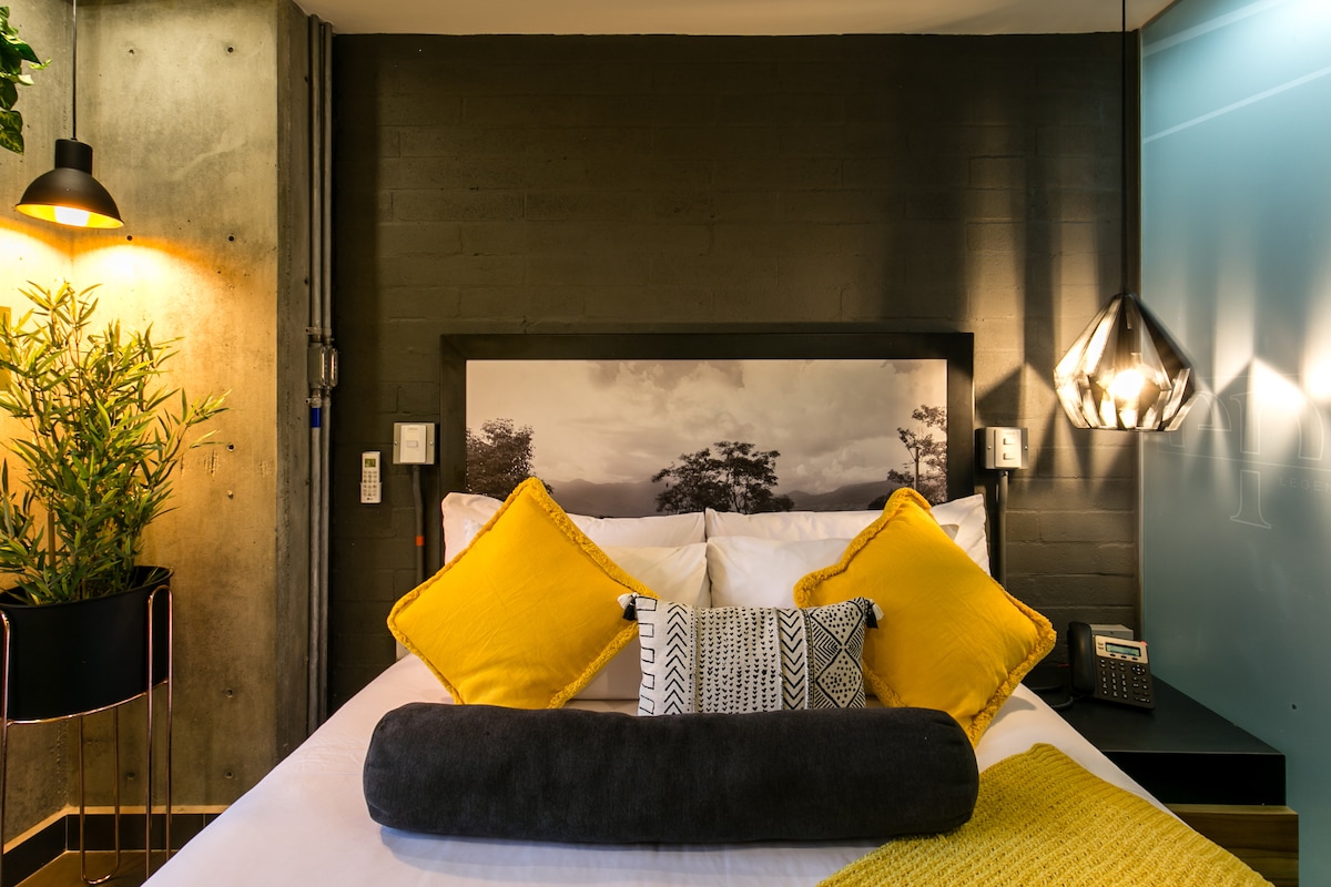 A modern sleeping area features a spacious bed adorned with yellow and patterned pillows. A textured wall serves as a backdrop, while a low plant stand adds greenery. Soft lighting is provided by a pendant light and a lamp, enhancing the contemporary design.