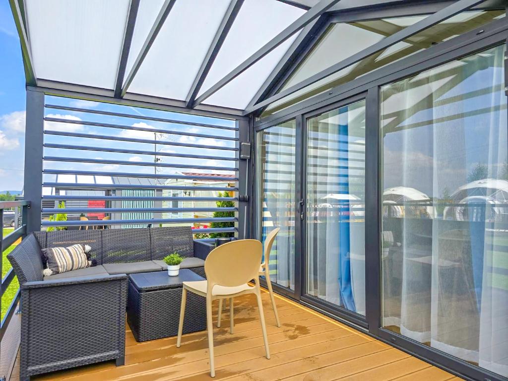 A covered terrace features a modern seating arrangement, including a dark wicker sofa with a small table. Light-colored chairs are positioned nearby. Transparent curtains are drawn, allowing natural light to filter through, while potted greenery enhances the tranquil setting.