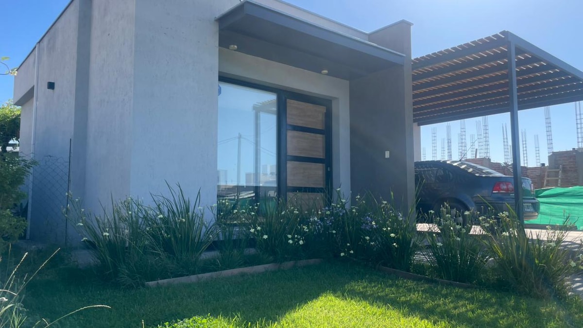 A modern exterior of a single-story house is presented, featuring a large glass door framed by greenery. A carport is visible to the right, providing shaded parking. The well-maintained lawn showcases lush grass and ornamental plants, enhancing the welcoming appearance of the home.