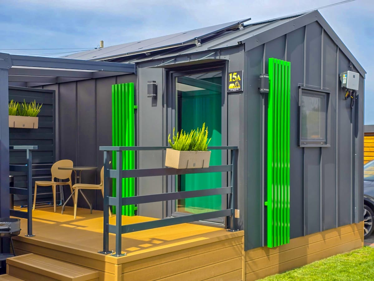 A modern cabin exterior features a wooden deck leading to a doorway framed by vibrant green accents. Comfortable seating is positioned on the deck, and a small planter is mounted beside the entryway, adding a touch of greenery to the structure.