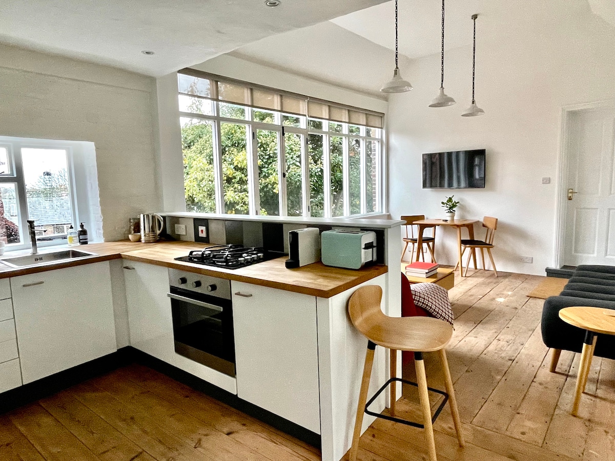 The kitchen features modern cabinetry with a wood countertop, including a gas stove and a microwave. Natural light floods in through expansive windows, illuminating the open living space which contains a dining table and comfortable seating. A TV is mounted on the wall.