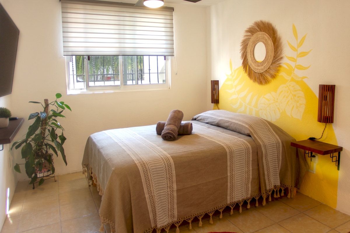 A spacious sleeping area features a stylish bed adorned with a neutral-toned blanket. Two wall-mounted lamps provide soft lighting, and a decorative mirror adds an elegant touch. A green plant sits in the corner, complementing the warm, inviting color scheme.