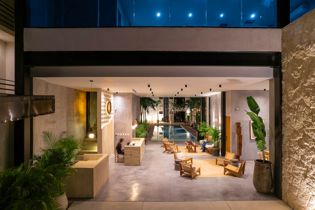 An open communal space features a modern design, showcasing warm wood seating and stylish light fixtures. A central pool stretches through the area, surrounded by lush greenery. The ambiance reflects a harmonious blend of indoor and outdoor living, all under an expansive ceiling.