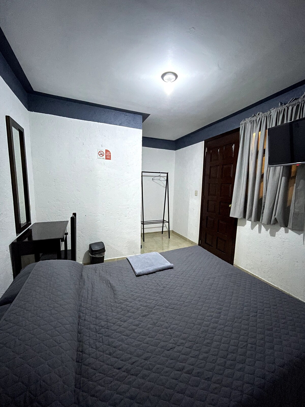 A cozy sleeping area features a textured gray bedspread draped over a spacious bed. A mirror and a small writing desk are positioned against the wall. A wardrobe stand is included, with light filtering through partially closed curtains, creating a calm atmosphere.