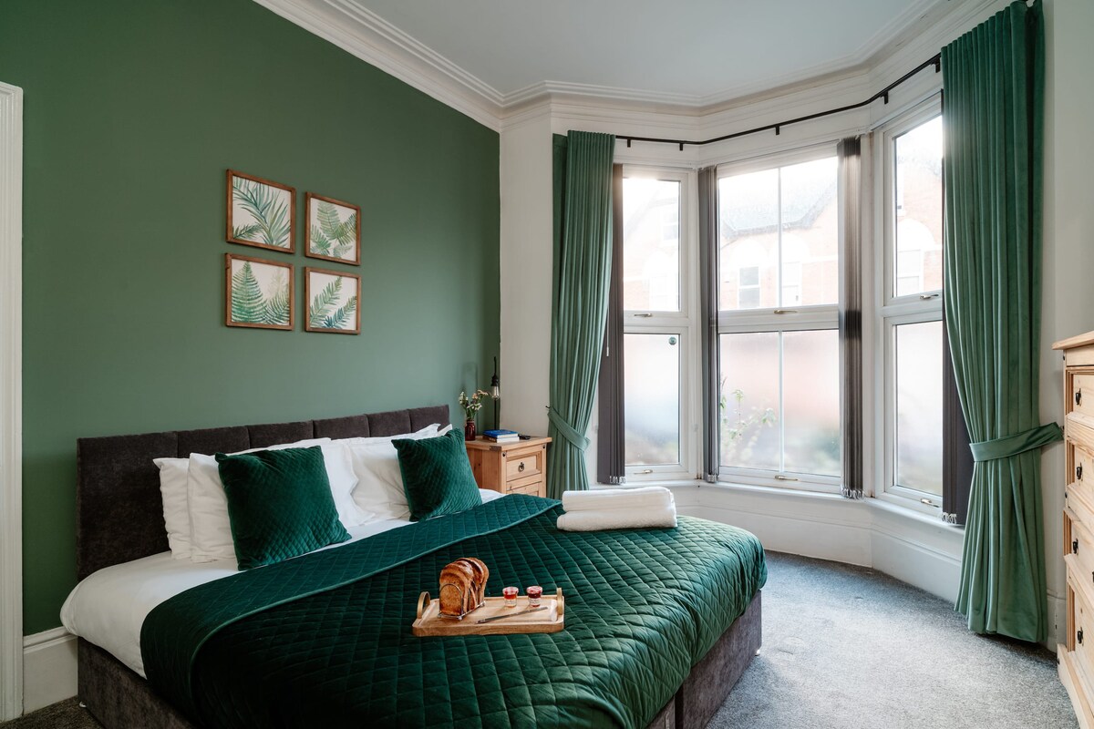 A spacious bedroom features a large bed with a dark quilt and plush pillows. Green curtains frame the bay windows, providing ample natural light. Wall art with plant motifs adds a touch of style, while a wooden dresser complements the decor.